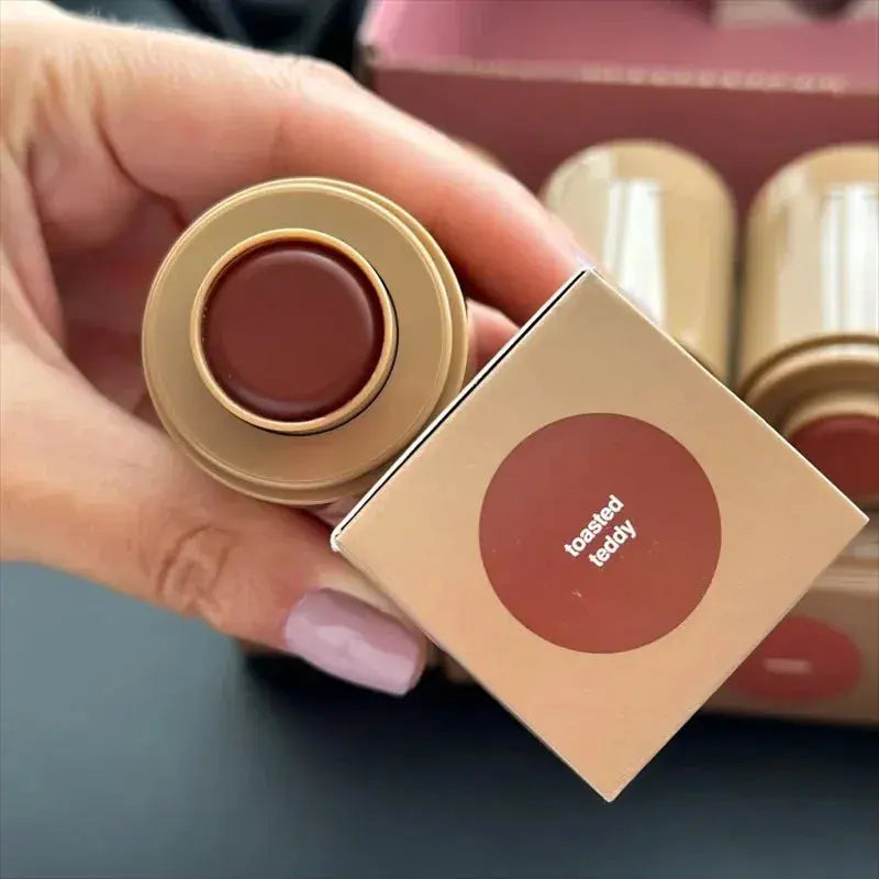 Lip glaze tin with dark brown product and circular label; hand holding the lid